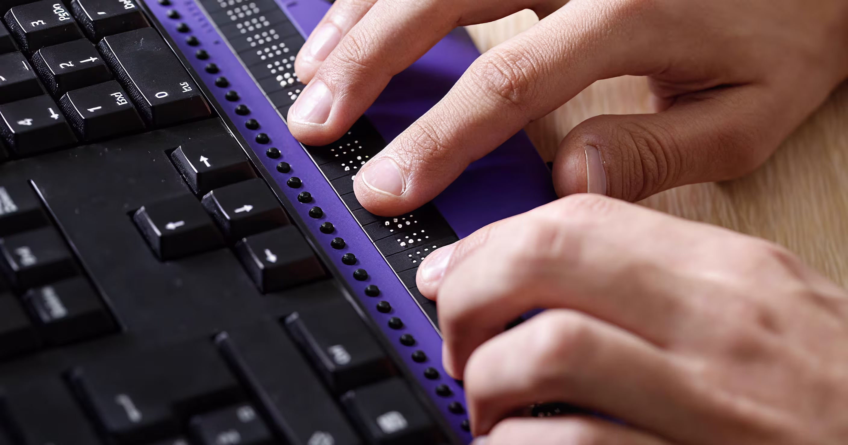 A close-up shot of a person's hands reading a purple braille display device that is connected to a black computer keyboard.