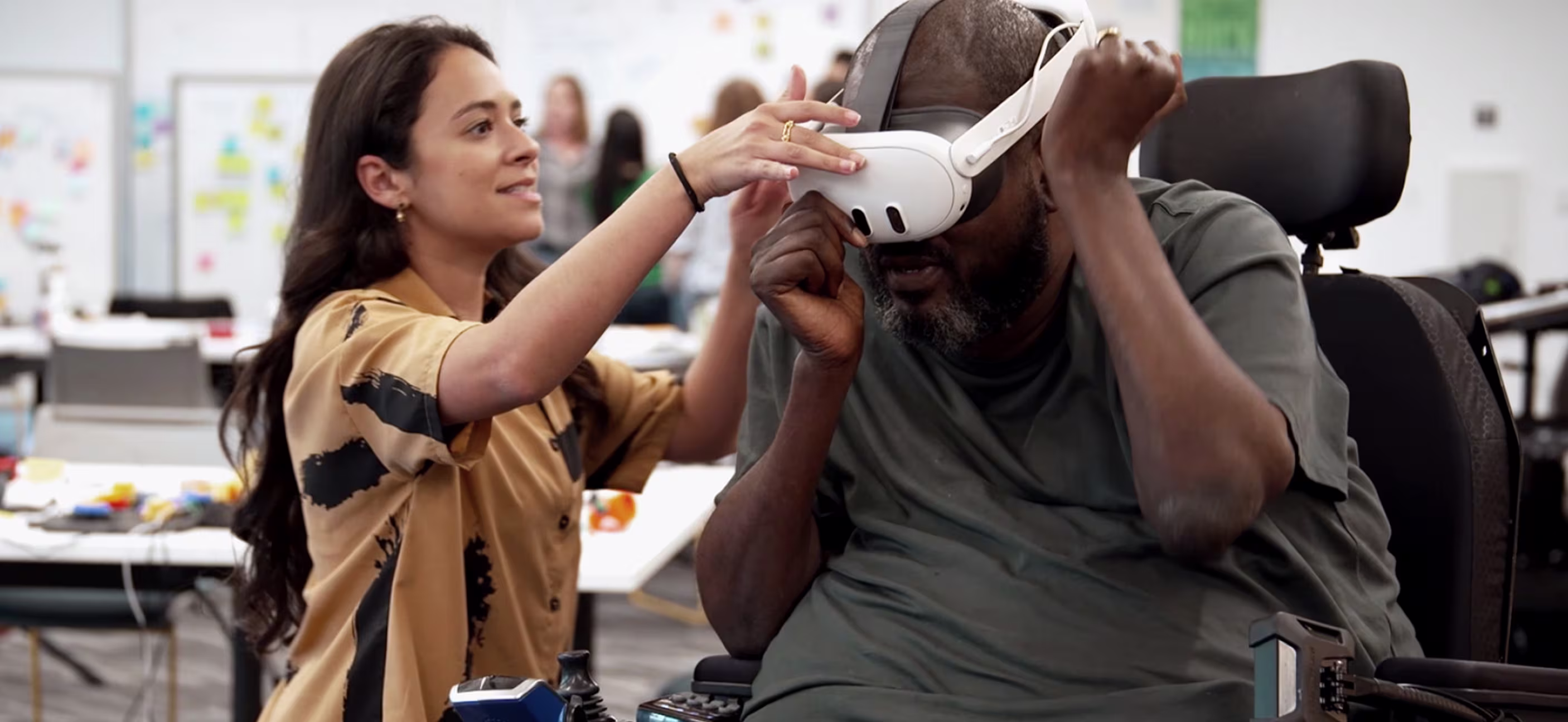 A woman with long dark hair, wearing a patterned shirt, assists a Black man in a power wheelchair with putting on a white VR headset.