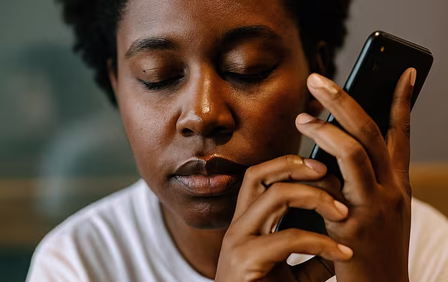 A close-up portrait of a Black person with short, dark, curly hair, holding a black smartphone to their left ear. Their eyes are closed, and their expression appears somber or contemplative.