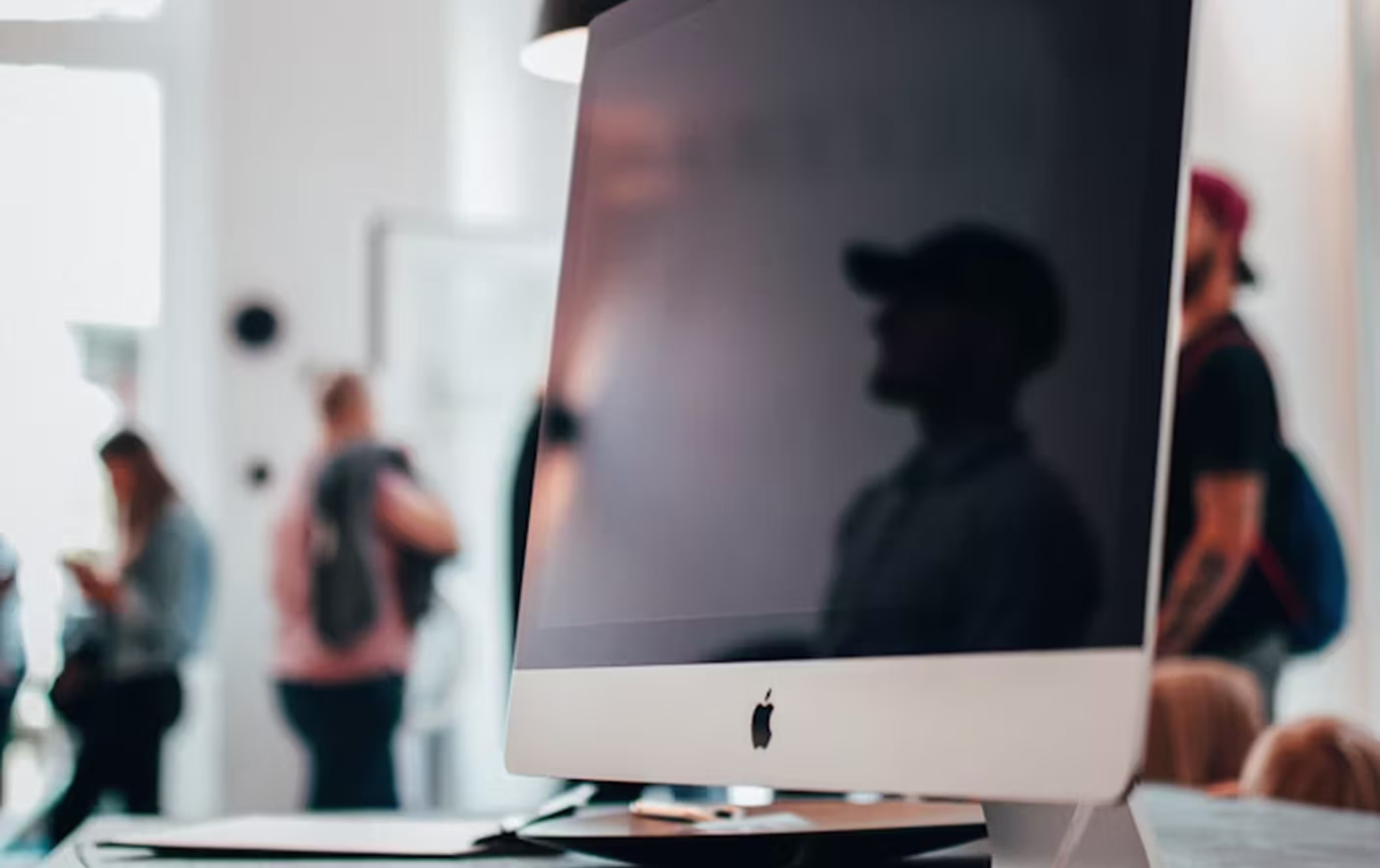 A modern office workspace featuring an Apple iMac monitor with a dark reflective screen, set against a blurred office background with warm lighting